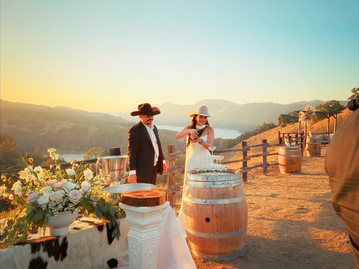 A couple in cowboy hats on a hilltop as she pours champagne into a glass tower, with mountains and a lake behind them