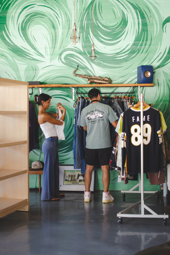 Two customers browsing vintage clothing racks against a green mural